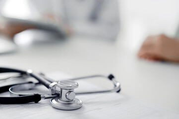 Doctor and patient are sitting and discussing something at the desk in the clinic office. The focus is on the stethoscope lying on the table, close up. Perfect medical service and medicine concept