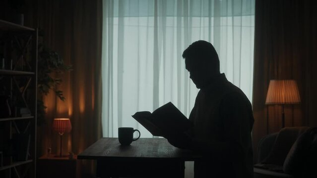 A man reads a book sitting at a table with a cup of coffee, tea in the room. Dark silhouette of a man with an open book in his hand, reading, turning pages, drinking coffee. Slow motion.