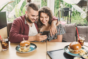 Happy young couple using  smartphone at burger pub restaurant. Young people having lunch break at cafe bar. Lifestyle concept with guy and girl hanging out on weekend day and having date