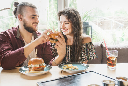 young couple sits in cafe and eats burgers with cola. Man and woman have a great time and relax enjoy food in cafe. Trendy wearing boy and girl have lunch. Teenager date