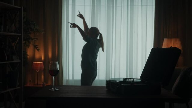 Vintage record player with a rotating vinyl record and a glass of red wine on the table close up. Carefree woman dances cheerfully alone. Dancing woman silhouette in the living room. Slow motion.