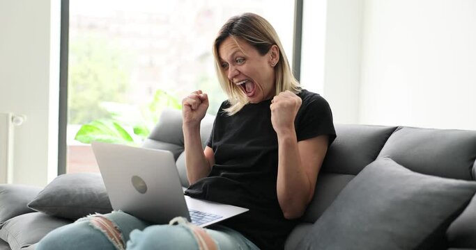 Euphoric woman winner celebrating victory triumph sits at home on sofa with laptop computer. Girl wins online fortune feels excited gets new job opportunity