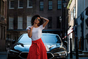 Black curly woman posing in front of luxury car.
