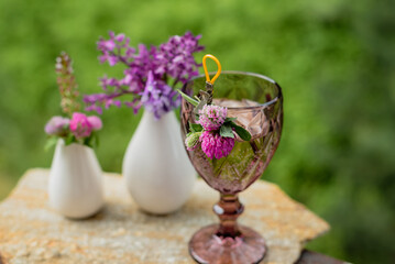 A glass with a drink and flowers in a white vase on the terrace against the backdrop of a green garden