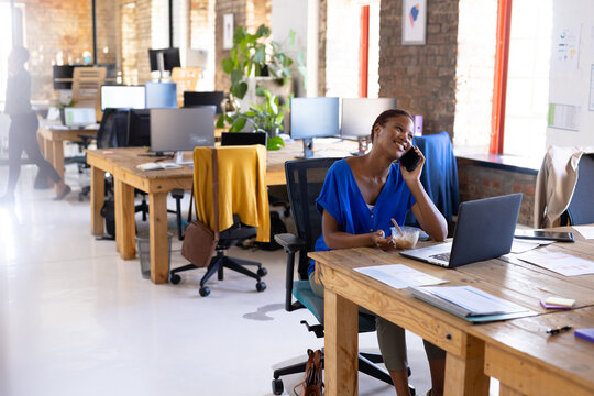 Happy african american casual businesswoman talking on smartphone and having lunch in office
