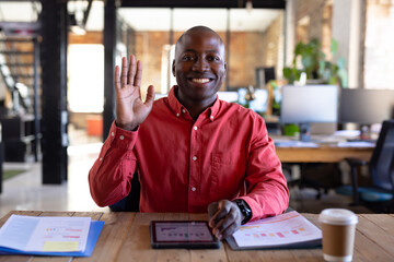 Portrait of happy african american casual businessman having video call and waving hand in office