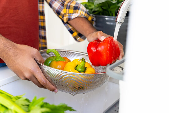 Midsection of biracial man rinsing peppers in colander at sink in sunny kitchen