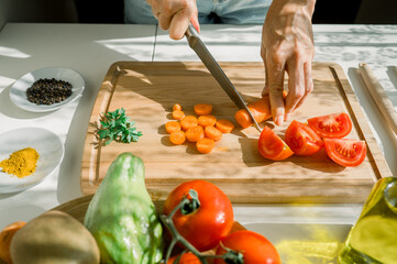 High angle of crop anonymous woman cutting carrot on wooden chopping board near tomatoes and...