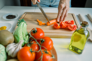 High angle of crop anonymous woman slicing fresh carrot on cutting board while preparing salad with tomatoes and parsley in kitchen