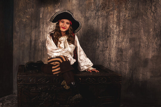 Portrait of smiling pirate sitting on chest in underground room or hold of pirates ship, playful looking at camera. Charming child girl actress in corsair image. Theatre perform concept. Copy ad space