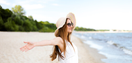 Happy smiling woman in free happiness bliss on ocean beach standing with a hat, sunglasses, and open hands. Portrait of a multicultural female model in white summer dress enjoying nature during travel