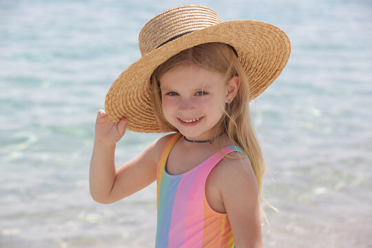 Outdoor portrait of fashionable little girl wearing straw hat on the beach with beautiful blue sea behind, summer vacation fashion and sun protection concept
