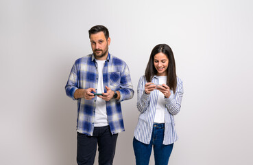 Young boyfriend with attitude looking at camera while playing video game with smiling girlfriend over smart phones. Couple gaming with cellphones and standing on isolated background