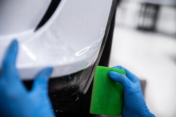 Car detailing studio employee, a wrapper, is protecting the bumper of a white car with a colorless...