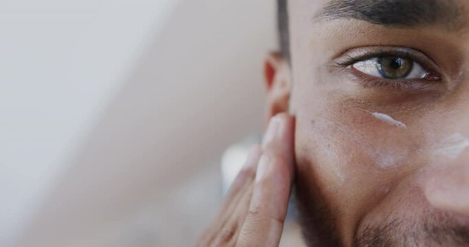 Close up of biracial man looking in mirror applying face cream under eyes in bathroom, slow motion