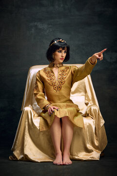 Portrait of beautiful young woman in image of Cleopatra, queen in elegant gold dress sitting and commanding over dark vintage background. Concept of antique culture, history, comparison of eras, art