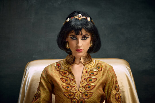 Close-up portrait of attractive, serious young woman in image of queen, Cleopatra looking at camera against dark vintage background. Concept of antique culture, history, comparison of eras, art, ad