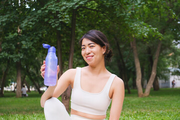 a sportif asian woman is holding bottle of water and sitting on yoga mat in park outdoor.