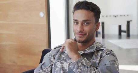 Portrait of happy biracial male soldier in uniform sitting in armchair at home smiling, slow motion