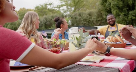 Diverse group of friends holding hands and saying prayer at dinner table in garden, slow motion - Powered by Adobe
