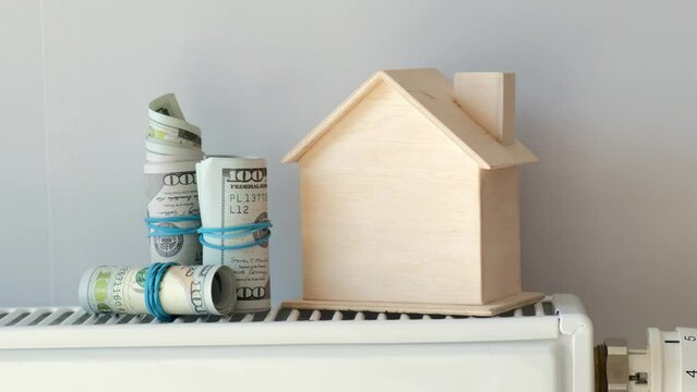 Dollar bills and a miniature house on the background of a radiator close-up. Concept of energy crisis, high bills, broken heating system, economy and saving money on monthly utility payments.