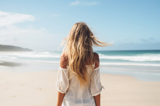 The back view of a young stylish woman walking along a pristine sandy beach, her long blond hair flowing in the breeze Generative AI