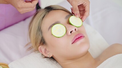 Obraz na płótnie Canvas Close up face of beautiful woman lying on spa bed while getting masking eyes with fresh cucumber slice. Therapist putting fresh cucumber slice on eyes of client woman. Skin care concept
