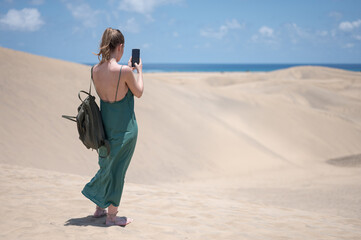Blond young girl in green dress and backpack in the dunes of the beach taking photos with the smartphone