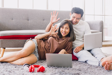 Beautiful young couple relaxing and smiling on a couch at home and making a video call to friends. Asian couple say hi to their family while using laptop in a beautiful living room.