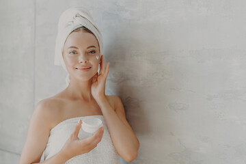 Lovely model applies moisturizer, holds cream jar, beauty routine, towel wrapped, poses against...