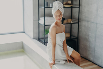 Relaxed woman with healthy skin smiles, towel-wrapped, uses cream, enjoys beauty routine, takes shower. Beauty, grooming concept.