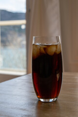 A tall clear pint glass of dark soda with frozen ice cubes floating on the top of the glass. The soft drink is a carbonated nonalcoholic soda. There are bubbles and fizzy in the glass sitting on table