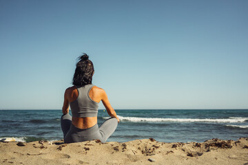Fototapeta na wymiar a beautiful athletic brunette girl in a gray tracksuit is engaged in meditation sitting on the sand against the backdrop of the sea
