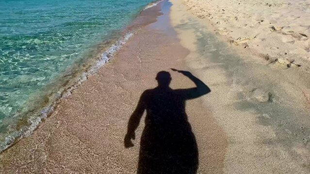 Silhouette shadow of man taking off sunglasses and walking on sandy beach along turquoise sea water shoreline