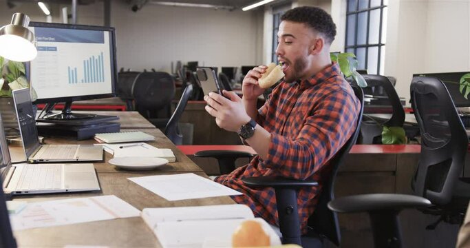 Happy biracial casual businessman sitting at desk, eating sandwich and using smartphone, slow motion