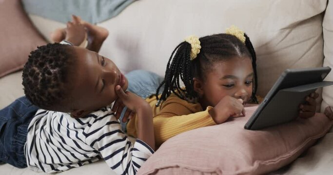 Happy african american brother and sister using tablet relaxing on sofa at home, slow motion