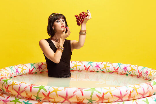 Beautiful young woman in image of Cleopatra sitting in swimming pool with milk and eating grapes against yellow studio background. Concept of antique culture, history, comparison of eras, art, beauty