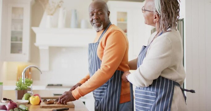 Happy african american senior couple in aprons preparing meal in kitchen, slow motion