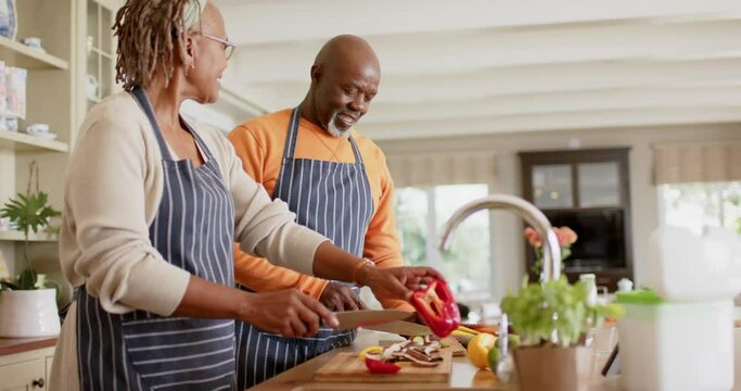 Happy african american senior couple in aprons preparing meal in kitchen, slow motion