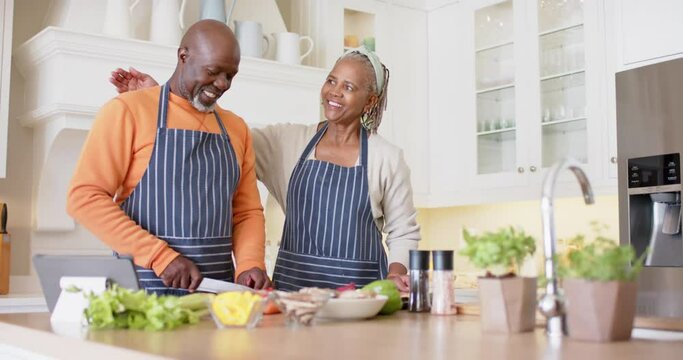 African american senior couple in aprons preparing meal using tablet in kitchen, slow motion