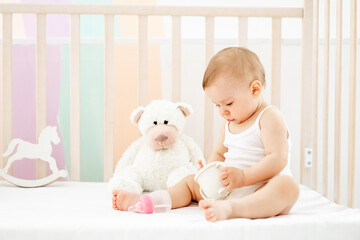 a little baby girl with a bottle or a drinking cup in her hands in a crib at home in a children's room in a white bodysuit smiling or laughing, cute funny baby, lifestyle