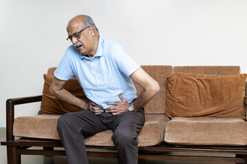 Senior men holding his stomach feeling pain while sitting on the sofa at home. Asian man having...