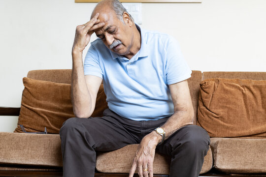 Tired, depressed senior man sitting on couch in living room feeling hurt and lonely. Aged, white-haired man touching forehead suffering from severe headache or recalling bad memories