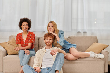 polygamy concept, diversity in relationships and culture, non monogamy, happy redhead man and multicultural women looking at camera in living room, lovers, acceptance, open relationship, banner