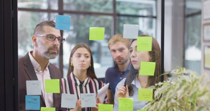 Work colleagues discussing startup project ideas. Team of caucasian business people standing near glass wall with sticky notes. Scrum methodology with colorful stickers. Backward camera movement.