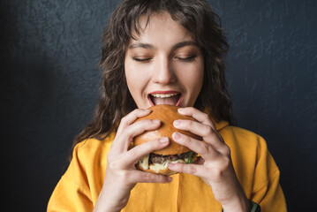 Attractive girl in a yellow hoodie eats a burger on a dark blue background closeup. Young woman...