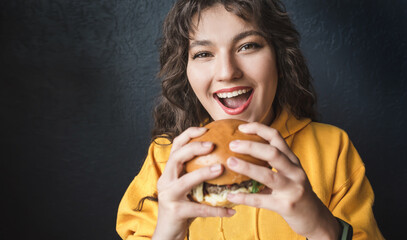 Attractive girl in a yellow hoodie eats a burger on a dark blue background closeup. Young woman...