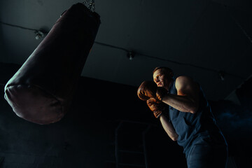 Male boxer in boxing gloves training on punching bag in dark gym.
