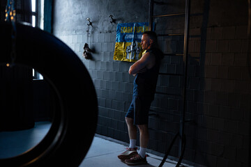 Obraz na płótnie Canvas Athlete resting in dark gym after exercising against background of Ukrainian flag with signatures of Ukrainian warriors and inscription.