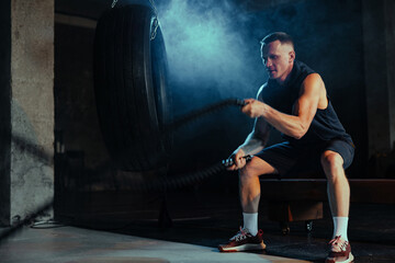 Strong muscular man exercising in dark gym with fighting ropes.